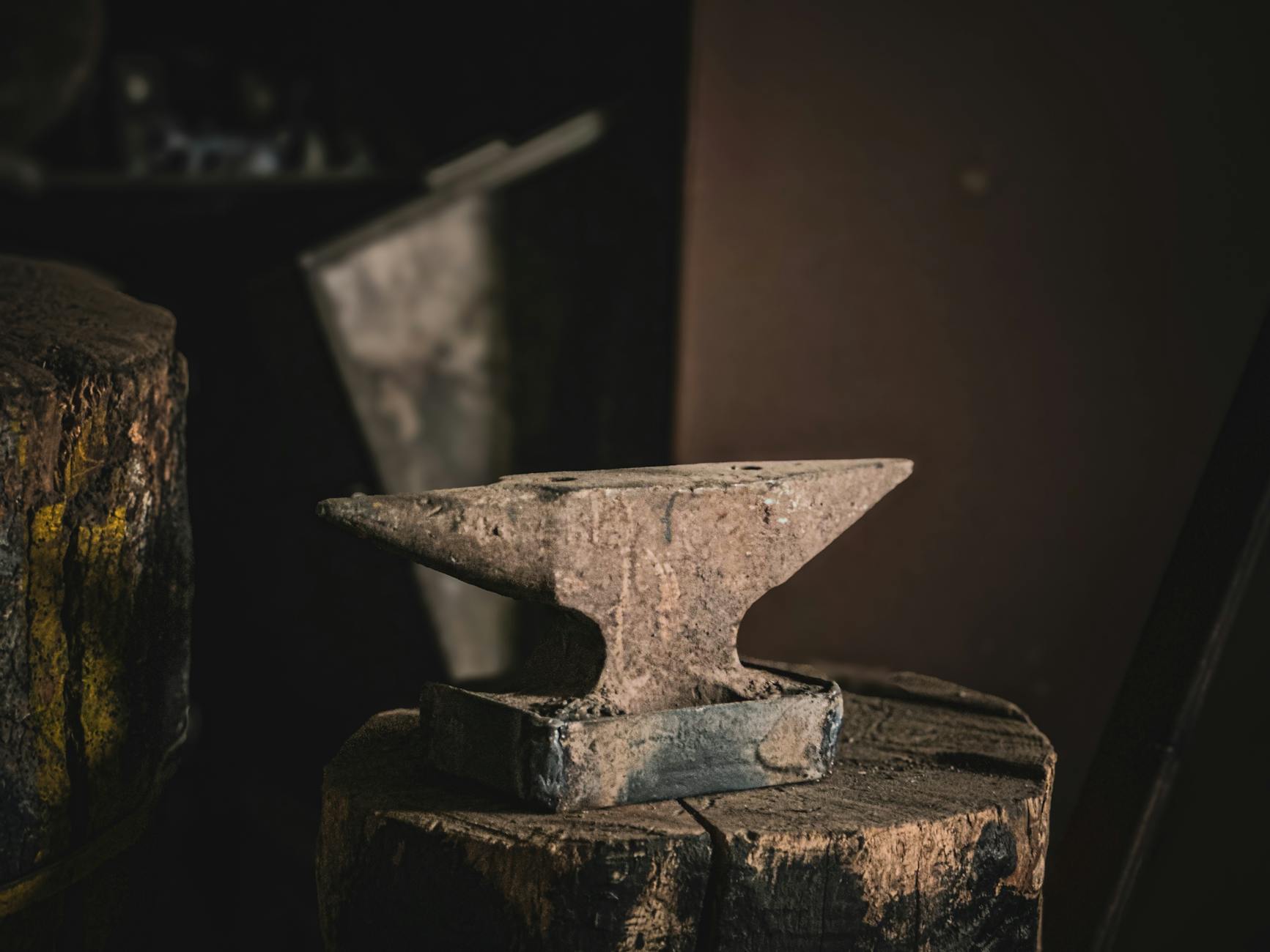 A rustic anvil sits atop a wooden stump in a dimly lit workshop, suggesting traditional blacksmithing.